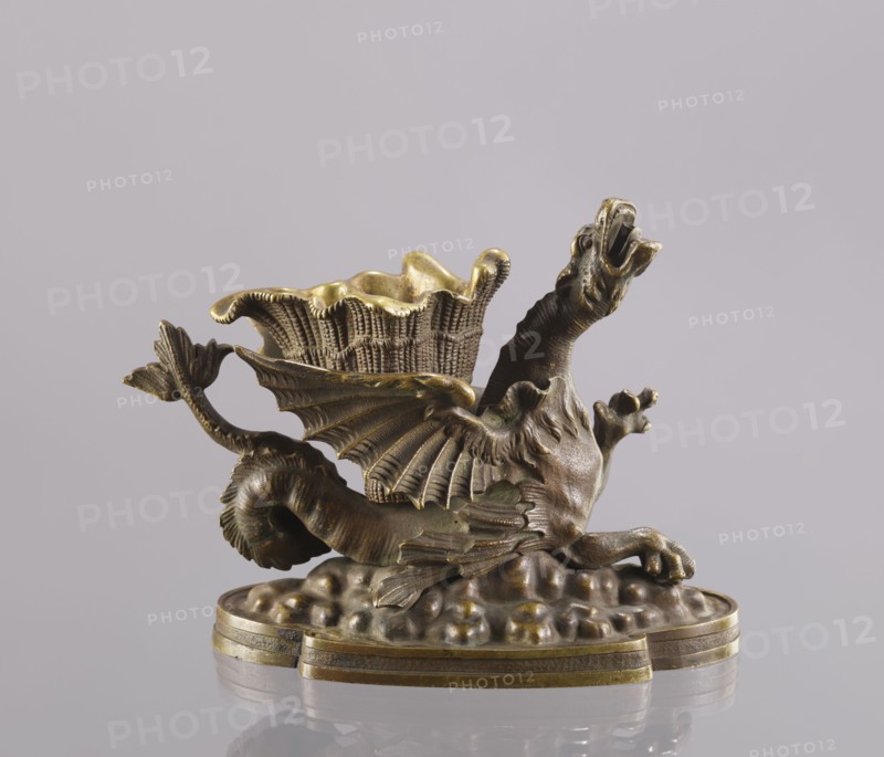 Spirit lamp, In the form of a two-winged dragon on a four-leaf oval base. Between the wings is a container; in the mouth is a hole for the wick, and on the side is a hole for pouring spirit. According to information in Szewczykowski’s catalogue, this is an oil lamp and a very old one. Due to the relatively large distance from the bottom of the container to the mouth, the lamp-burner cannot be fired with oil, which has poor capillarity. Most likely the lamp was fired with spirit, and the container between the wings was used to hold cigarettes. 2nd half of the 19th century, Polesie. Lamp; height 15.4 cm, width 20.8 cm, depth 10.5 cm., A small sculptural object depicting a mythical creature with a long serpentine body, outstretched wings with ribbed membranes, a raised neck and open mouth, and clawed forelimbs, all rendered in a metallic material with aged golden and bronze tones; the creature reclines on a textured rounded base that has a layered rim, and a basketlike cup with a ruffled ...