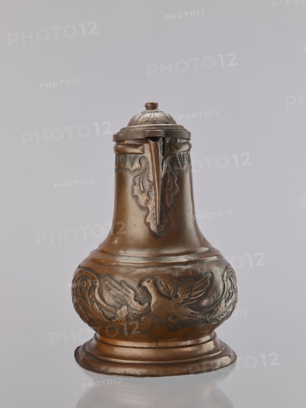 Jug. On a circular, stepped base, the body bulges in the lower part, decorated with a repoussé leafy twig and, at the rim, an eagle with spread wings. Above the two stepped areas is a neck with a spout. Repoussé leaves under the rim and along the rim. The hemispherical handle, fastened with rivets; the lid is hinged with a finger grip, arched, decorated with a leafy rosette and topped with a ball. Maker: Roman Szewczykowski 1849–1901. 1st half of the 18th century. Poland. Vessel. Height 39.5 cm, width 23.9 cm, depth 28.4 cm., A single metal pitcher with a domed lid and a small finial sits against a plain light gray background, showing a warm brown and bronze surface with darker patina in recessed areas. The vessel has a bulbous lower body that tapers into a narrower neck and a flared base, with decorative raised relief carving around the midsection depicting a bird with outstretched wings amid stylized foliage and scrolling motifs. The neck and upper shoulder feature leaf-like relief p...