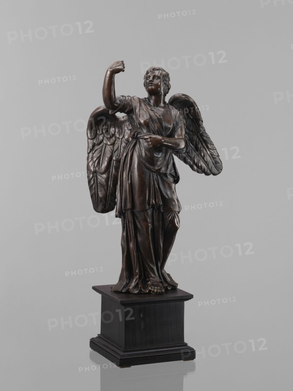 Statue - Angel, Figure of a standing angel with his right hand raised, the other raised to chest height, which originally contained a cross or an element of the Arma Christi. It is a pendant to another angel. Square wooden base with profiles on the upper and lower edges. Renaissance style, 16th century, sculpture; height 45 cm, width 12 cm, depth 12.5 cm., A dark brown statue of a winged figure stands on a black rectangular base against a pale gray background, showing sculpted drapery over the body, detailed feathered wings extending behind the shoulders, one arm raised and the other bent across the torso, with visible facial features, curly hair, and bare feet peeking from beneath flowing garments.