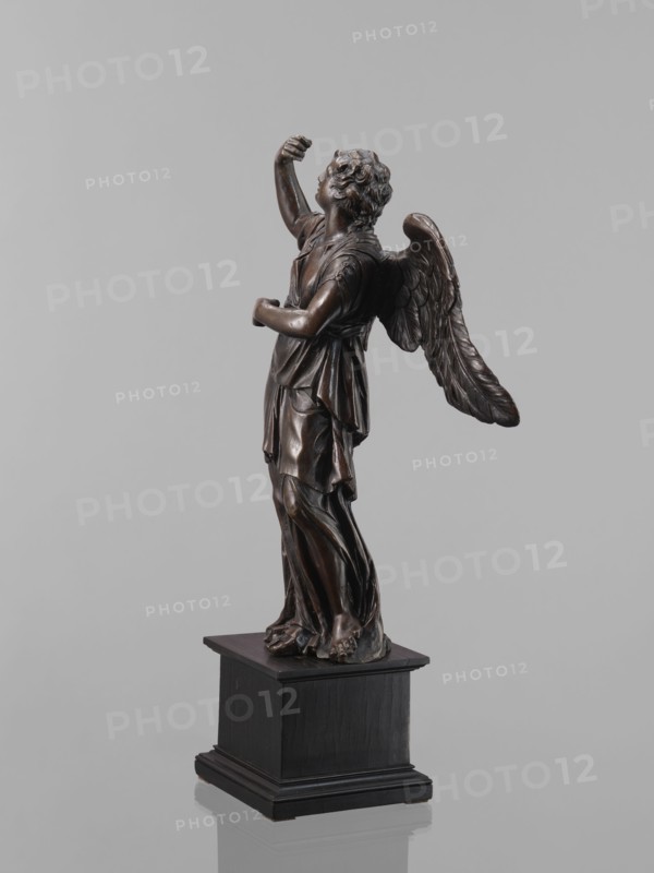 Statue - Angel, Renaissance, 16th century, Figure of a standing angel with his right hand raised, the other raised to chest height, which originally contained a cross or an element of the Arma Christi. It is a pendant to the latter. Square wooden base with profiles on the upper and lower edges, a secondary one. Sculpture, height 45 cm, width 12 cm, depth 12.5 cm., A dark brown metallic statue of a winged figure stands on a black square pedestal against a light gray background, showing a draped robe with flowing folds, curly hair, raised arm and an upward gaze, with textured feathered wings extending behind and bare feet visible at the base.