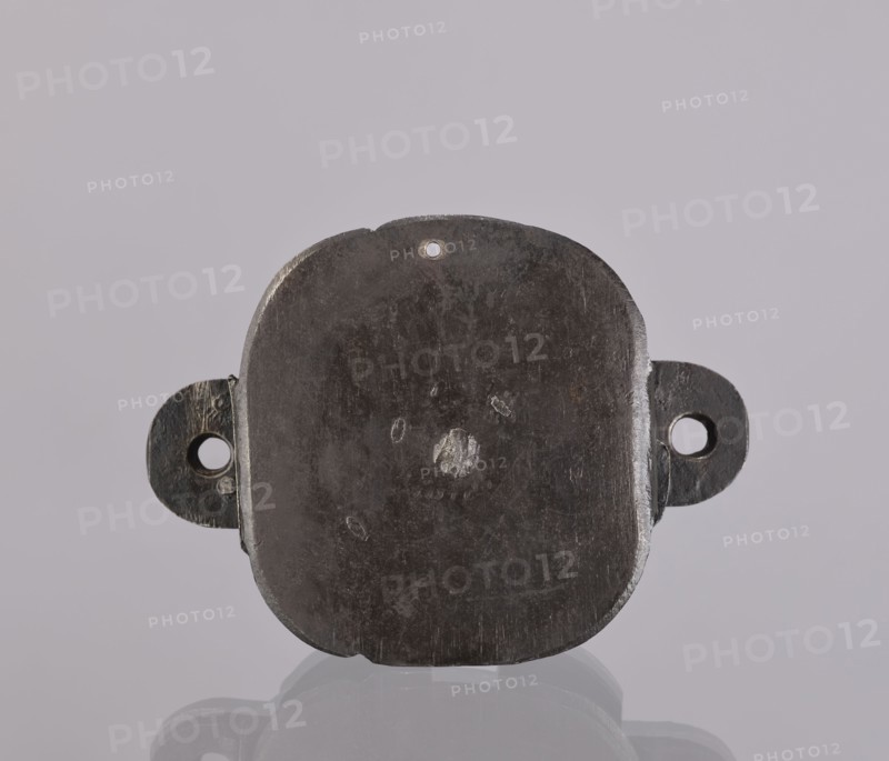 Lock register. The register on the bottom plate is in the form of an irregular oval, with two semicircular projections with mounting holes that turn into flat supports for the cover in the form of a rectangle with rounded corners. Inside the key rod is a guide made of double sheet metal; on both sides of it are single horizontal slats, and two on the bottom plate of the register. 19th century, Germany. Lock. Height 11.7 cm; width 16.3 cm; depth 5.3 cm., A dark gray, roughly square metal object with rounded corners and a worn, scratched surface sits centered against a light gray background; it has a small round hole near the top edge and a few shallow dents and abrasions on its face, with a pair of rounded side tabs extending left and right, each featuring a circular opening; the object casts a subtle reflection and shadow on the glossy surface beneath it.