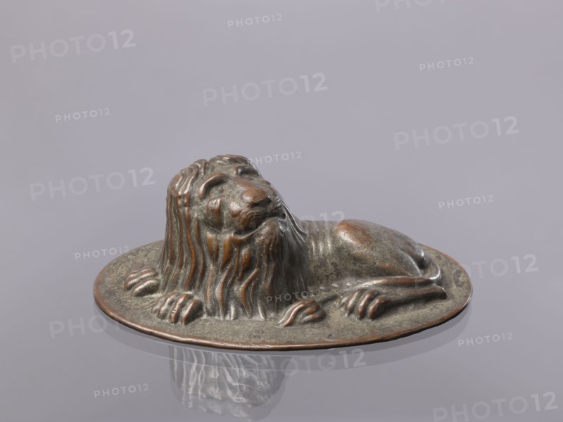 Jelly mold, repoussé copper sheet in the form of a lying lion; raised head allows contents to be removed intact., described variously as a button or jelly mold. 18th–19th century, possibly Germany. Vessel; height 5.4 cm, width 15.5 cm, depth 9.8 cm., A small sculpted lion figure resting on an oval base, showing a reclining pose with its head raised and mane flowing down its neck and chest; the surface of the sculpture appears metallic with a mix of greenish patina and brownish-bronze highlights, and the base shares the same coloration; the background is a smooth, light grayish-lavender surface that reflects the sculpture faintly.