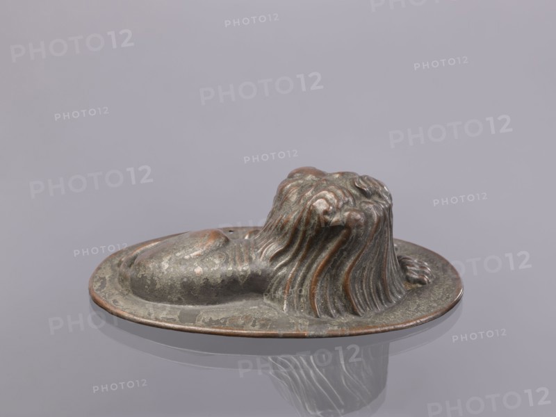 Jelly mold, repoussé copper sheet in the form of a lying lion with raised head allowing contents to be removed intact. Probably 18th–19th century, Germany, vessel; height 5.4 cm, width 15.5 cm, depth 9.8 cm., A small sculpted object resembling a reclining animal with flowing mane-like ridges sits on an oval base; the surface shows a weathered metallic finish with greenish-gray and brownish-copper tones, with patches of darker and lighter areas and a subtle reflective sheen on the base, all set against a smooth pale gray background.