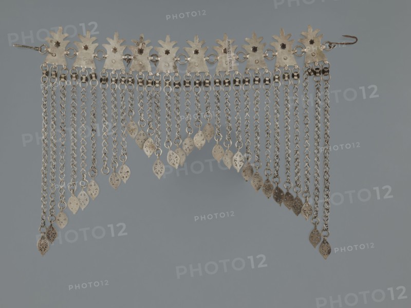 Necklace, 19th century, Middle East, Islamic art; pendants, body and garment ornament, female ornament; geometric ornament, buttons ornament, fashion accessory; jewelry; height 22.0 cm, width 18.0 cm, weight 131 g, A metallic decorative piece with a row of leaf-shaped plates along the top connected by small rings, from which hang a group of chain strands of varying lengths ending in small pierced leaf-shaped pendants; the metal appears silvery with areas of darker patina and spots of brownish discoloration, all set against a smooth muted gray background.