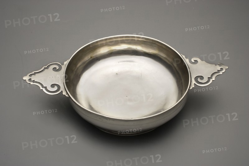 Écuelle by Christian Lieberkühn fl. 1703–1733, goldsmith. Hidden on the bottom: city mark not recorded in the literature, maker's mark Szeffler, pp. 58–59, no. 90. Circular, bulging, smooth vessel on a low base with two horizontal handles with openwork. Vessels of this type, appearing already in the 13th–14th centuries, were used for drinking broth and soups; they usually had a plate–stand and a cover topped with a handle or three legs, in the latter case serving as a stand. Unpublished. Berlin, Germany. Vessel; height 7 cm, width 25 cm, depth 15.5 cm, weight 333 g., A shallow round metal bowl with a smooth reflective interior and ornate flat handles extending from opposite sides, set on a flat gray surface; the bowl and handles appear silver in color with subtle darker and warmer metallic reflections, and the background is various shades of gray.