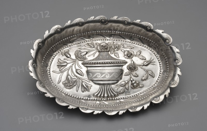 Platter by Johann Jacob Haase II active from 1759, died 1775, ca. 1780. Oval cake stand with a mirror decorated in relief with a covered vase topped by a bouquet of carnations, tulips, and roses; surrounded by a band of chien courant running wave motif enclosed in pearling, above it a rooster's comb; irregularly cut edge with a repeated running-wave motif. At the bottom a dotted inscription with the date MM & Fr.ACM / Ao 1782 4 Junii. Note: discrepancy between the goldsmith’s recorded activity died 1775 and the 1782 hallmark of Ellerholtz; studies do not confirm whether the widow or son continued the workshop. Silver; vessel. Measurements: height 2.2 cm, width 21.9 cm, depth 15.2 cm, weight 159 g. Made in Gdansk, Royal Prussia, Poland., A shallow decorative oval metal dish with an ornate scalloped rim and embossed floral and urn motifs, shown against a smooth gray background; the dish displays polished and matte areas with visible texture, a raised beaded inner border, stylized leaves ...