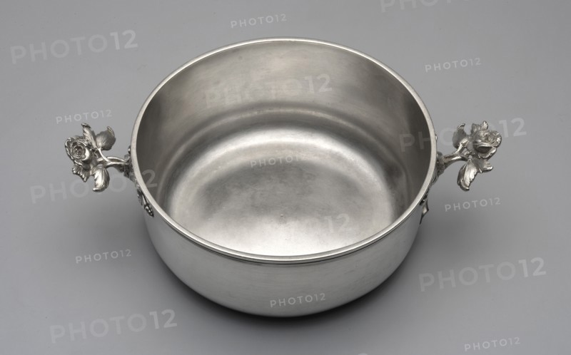 Vegetable pot, maker: Ephraim Wischcke fl. 1755–1799, goldsmith. Breischüssel type of vessel. Round vegetable pot with a flat bottom and profiled rim; handles formed as two plastic rose stems; no lid. Probably intended for serving boiled, chopped, or pureed vegetables hence called Breischüssel in German literature. Decoration: flower on stem rose. Date: 1770s. Place: Gdansk, Royal Prussia, Poland. Medium: vessel. Dimensions: height 7.4 cm, width 25 cm, depth 17.3 cm; weight 727 g., A metallic round bowl with a smooth interior and exterior surface sits on a flat background, showing a silver-gray sheen. The bowl has a simple rim and two decorative handles shaped like detailed flowers with leaves extending outward, also in silver-gray. The background is a uniform light gray. The overall visible colors are silver-gray and light gray.