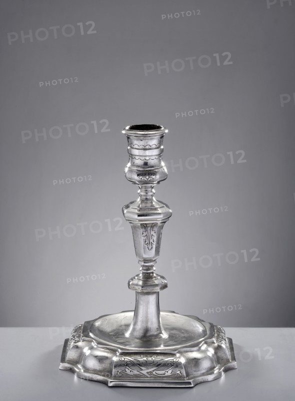 Single-candle candlestick by Hieronymus Holl II 1660–1732, goldsmith. On an octagonal foot with alternating straight sections, in the form of a half-cylinder with pincer and wavy decoration. The main surface is circular, slightly concave, from which the lower part of the shaft extends, terminating in an octagonal, flattened node. Above, the shaft is an octagonal baluster widening upwards; the sides are alternately smooth and engraved with acanthus leaves. Cylindrical profiled sleeve, expanded at upper and lower parts. Dated 1721–1730, made in Gdansk, Royal Prussia, Poland. Function: lighting. Height 14.5 cm; width 11.2 cm; weight 207 g., A single metallic candlestick stands centered on a plain surface against a neutral background, showing a shiny silver color with polished highlights and subtle darker tones in recessed areas; the candlestick has a flared base with decorative raised edges and engraved floral motifs, a tapered central stem with rounded and faceted sections, and a cup-lik...