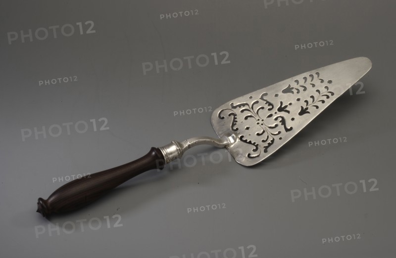 Cake server by Johann Christoph Conradi fl. ca. 1717–1742. Triangular with rounded corners, decorated with openwork in the form of plant tendrils and flower calyxes. The handle is curved, ending in a polygonal socket containing a wooden reconstructed handle. Typically this type of server is described in the literature as intended for fish, though the later typeification of shapes for fish, cakes, tarts, pâtés is noted. Dated 1717–1742, Berlin, Germany. Cutlery/table accessory. Height 22 cm; width 7.9 cm; weight 121 g., A metallic cake server with an ornate perforated triangular blade featuring decorative cutouts rests on a gray surface, attached to a curved metal neck and a dark brown wooden handle with a rounded end and subtle turned detailing; visible colors include silver, dark brown, and shades of gray.