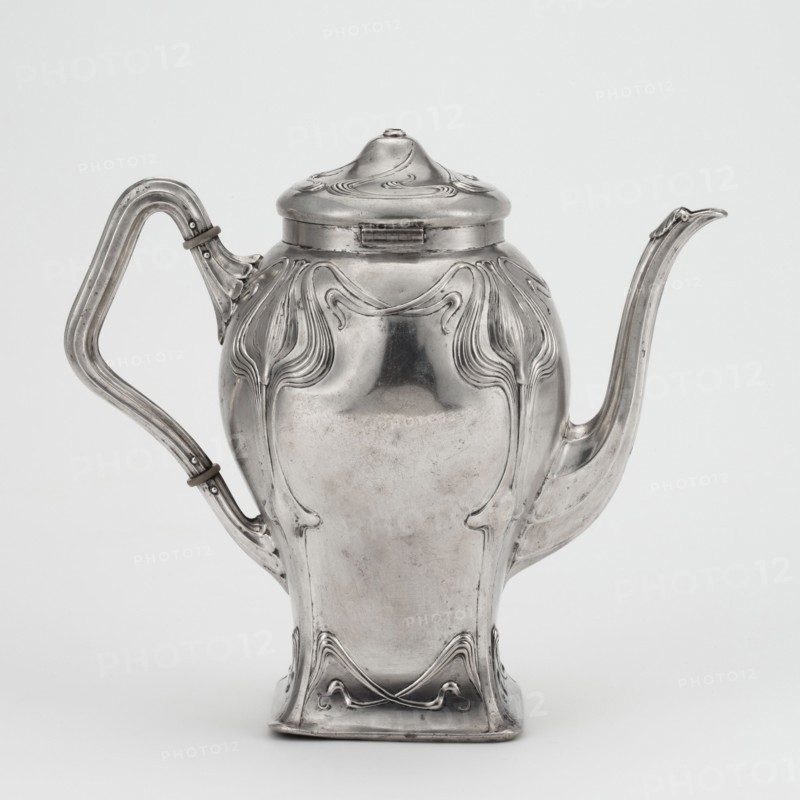 Art Nouveau coffee pot, Köberlin, Gebrüder Döbeln; goldsmith's manufactory; founded 1828 maker; Art Nouveau style; plant ornaments stylized plant twig; coffee pot on a square plan with slight narrowing in the lower section and a strong bulge in the upper section; S-shaped spout; handle broken twice; hinged lid, slightly bulging, with a broken handle; stylized twigs in the lower section terminating in flowers in the upper section; bone handle insulators; engraved on the bottom: F. und M. / von Pentz-Brandis / Mai 1902; circa 1900; Germany, Döbeln, Saxony; vessel; height 20.4 cm, width 23.5 cm, depth 11.5 cm; weight 589 g., A metal teapot with a shiny silver surface, featuring a rounded body that tapers to a square base and a domed lid, decorated with flowing raised ornamental lines and curved relief motifs; a sculpted spout extends from one side and a curved handle with a few metal bands is attached on the opposite side; the background is a plain pale gray, and the visible colors are si...