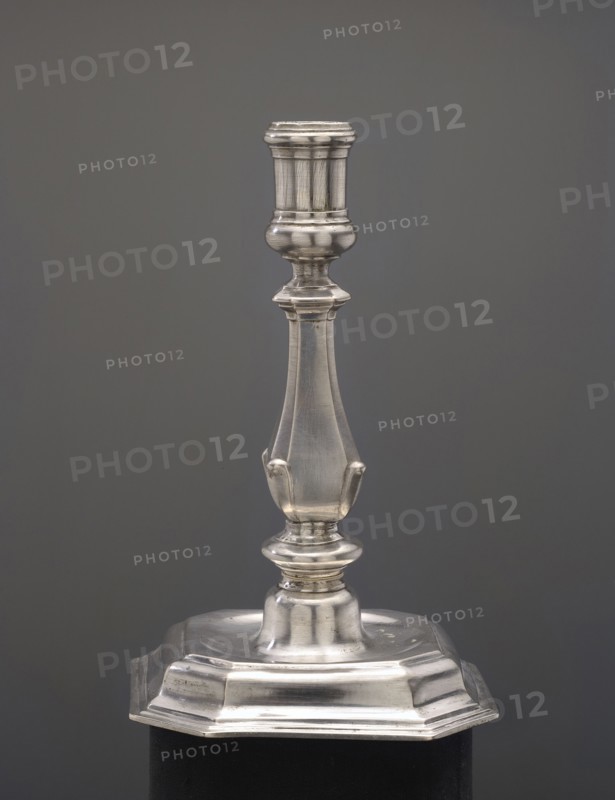 Candlestick, Spindler, Sebald fl. ca. 1716–1740, goldsmith. Candlestick on an octagonal profiled foot, baluster shaft narrowed towards the top, cylindrical candle socket. 1730s, Berlin, Germany. Candlestick lighting. Height 16.5 cm; width 10.3 cm; weight 292 g., A metallic candlestick with a polished silver finish stands against a smooth gray background; its design features a flared, ridged top leading to a bulbous neck, a tapered shaft with subtle curved panels and small projecting tabs, a rounded collar above a stepped, rounded base, and an octagonal lower platform with beveled edges resting on a dark support.