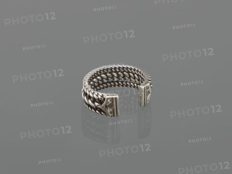 Bracelet; maker, 19th century; Middle East possibly; Islamic art; body ornament—female hand ornament; tugra motif; jewelry; materials unspecified; height 2.5 cm; width 7.0 cm; weight 170 g., A small metal ring with a braided design forming several intertwined rows, open at one side with two flat end caps that have textured surfaces; the ring and its reflection rest on a smooth surface. Visible colors include silver, gray, and tonal variations of those shades.