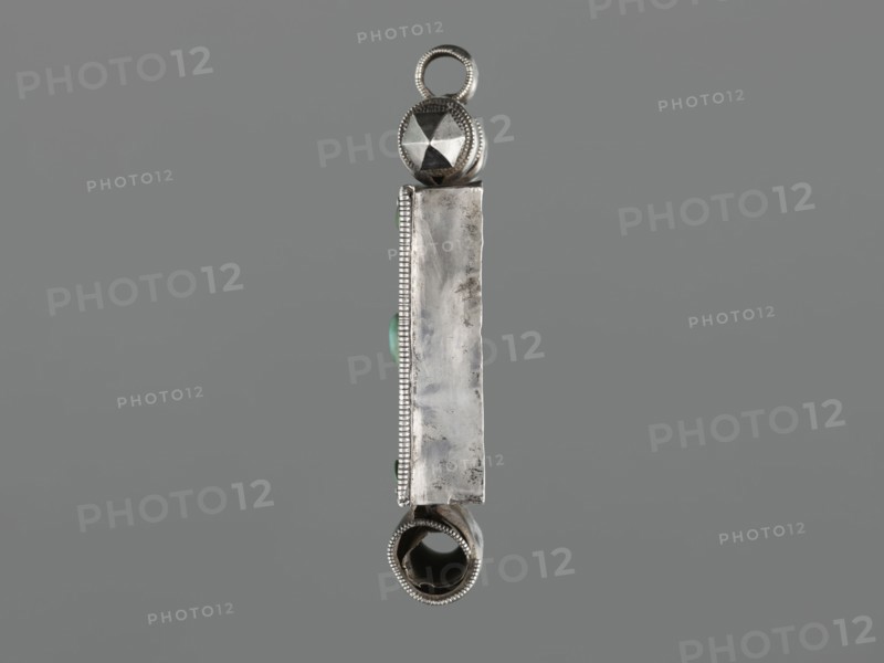 Ga'u box - fragment, plant twig, jewelry, Himalayan, Himalayan art, Tibetan Buddhist art, amulets, Tibetan culture, provenance unknown, 19th century, Tibet, varia, height 11.0 cm, width 7.5 cm, weight 125 g, A vertical silver metallic pendant photographed against a neutral gray background, with a rounded loop at the top and a hollow cylindrical element at the bottom, a faceted spherical bead near the top, an elongated rectangular centralplate with a slightly worn surface and beaded edging on one side, small rounded green stones set along the side, and visible textures and reflections on the metal showing areas of polish and subtle tarnish in colors including silver, gray, green, and dark metallic tones.
