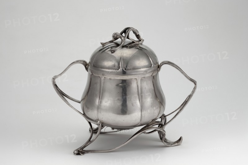 Art Nouveau sugar bowl, Art Nouveau style, plant twig motif, sugar vessel gilded inside, circular with an arched lid with a handle in the form of branches. Large twig-shaped handles extend into three tall supports of the body. Dated 1899–1907. Place of origin uncertain: Germany or Austria. Weight 410 g, height 15 cm, width 18 cm., A polished metal container with a rounded, bulbous body and a domed lid topped by an ornate looping handle sits on an elegant openwork stand; the container has two sweeping curved handles on opposite sides and the stand features sinuous, ribbon-like supports that curl at the ends. The surface is reflective and smooth with visible panels and subtle seams, showing highlights and soft shadows. The visible colors in the image are shades of silver and gray against a light gray background.