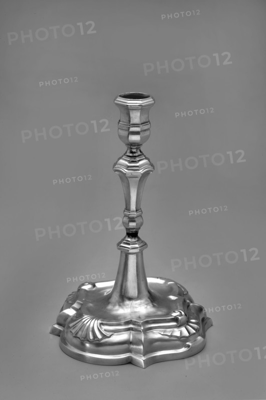 Candlestick by Johann Gottfried Schlaubitz fl. 1733–1771, goldsmith. Scrap - purchase, purchase provenance. On a quatrefoil foot, arched and profiled. In the center of the segments are alternating shells and volutes with a fan motif. From the slightly concave surface a conical lower part of the shaft protrudes, upon which a baluster with octagonal profiles is placed. Vase sleeve with octagonal profiles. Dated 1740s. Origin: Royal Prussia, Gdansk, Poland. Function: candlestick lighting. Dimensions: height 19.4 cm; width 13.1 cm; depth 13.2 cm. Weight: 295 g., A single metallic candlestick stands upright against a plain background, showing a polished reflective surface in shades of gray and silver. The base is ornate with curved, scalloped edges and flowing, embossed designs that catch highlights and shadows. The stem tapers upward with a series of decorative, faceted sections and rounded nodes, each reflecting light differently to create contrasts of brighter silver and darker gray. The...