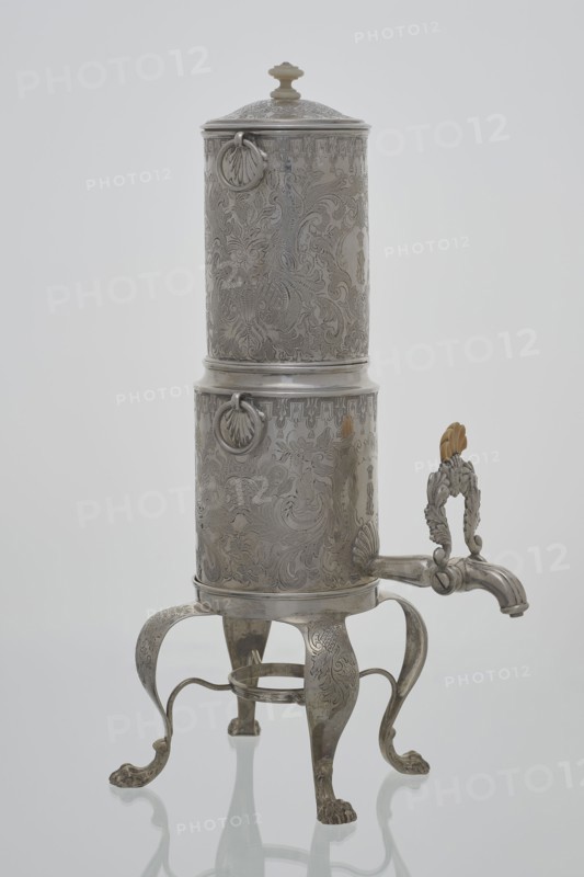 Coffee maker by Stephan Mayerhofer fl. 1818–1852, goldsmith; 1835, Vienna, Austria. Vessel with lion’s paw feet; height 35 cm, width 15 cm, depth 19 cm; weight 960 g., A tall ornate metal vessel with a cylindrical body and a domed lid sits on a decorative four-legged stand; the vessel and stand have intricate engraved floral and scrollwork patterns all over their surfaces, and there are a few circular ring handles attached near the top and middle of the cylinder. The stand's legs curve outward and end in clawed feet, and a curved spout extends from the lower portion of the cylinder with a decorative handle above it that features leaf-like motifs and a small carved element in a light tan color. The overall colors visible are silvery metal tones, light tan on the small carved element, and subtle reflections of white and soft gray from the background.