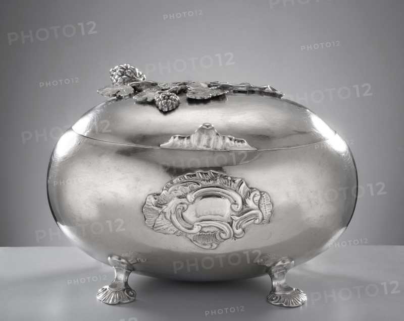 Rococo sugar bowl by Emmanuel I Ellerholtz fl. 1758–1800, goldsmith. Ovoid sugar bowl supported on four bent shell legs; front has a relief rocaille blind reserve. Hinged lid with a handle in the form of a rooster’s comb, topped with a modeled vine branch with clusters. Materials/ornaments: rocaille, vine, rooster’s comb. Style: Rococo. Date: 1768, Gdansk, Royal Prussia, Poland. Function: sugar vessel. Dimensions: height 10 cm, width 14.5 cm, depth 10 cm; weight 385 g. Provenance: purchase., A shiny metallic oval container with an ornate lid sits on a flat surface supported by a few short decorative feet; the surface of the container is reflective and smooth with embossed ornamental detailing on its front and floral-like embellishments on the lid. The colors visible are shades of silver and gray across the container and lid, with lighter silver highlights where light reflects and darker gray shadows on the underside and background. The background is a gradient of gray.