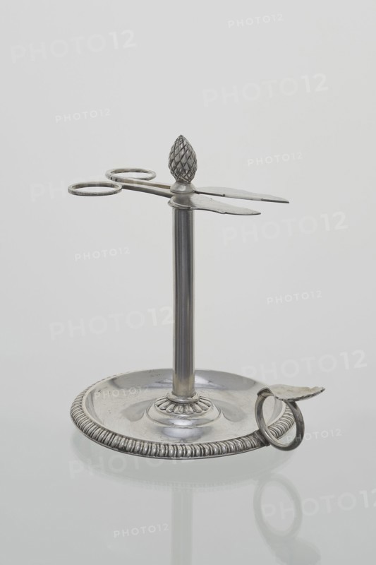 Scissor-shaped candlestick stoczek, Jan Radecki fl. 1815–1837, goldsmith. Candlestick for a thin candle that was serpentine-twisted around the stem; supported on a plate-shaped base edged with a half-cylinder and provided with a ring handle with thumb rest. Stem formed as a sleeve around which the thin candle twisted serpentine; sleeve topped with a pine-cone finial with a vice in the form of flat scissors with volute handles to extend the candle to desired length. 1815–1837, Poznan, Poland. Function: lighting. Height 14.5 cm; width 14 cm; depth 11 cm., A metallic stand with a circular base and a vertical central rod supporting a horizontal scissor-like element topped by a textured finial shaped like a pinecone; the base has a ridged outer rim and a small curved handle attached at the edge, and the object is shown against a plain light gray background. The visible colors are silver and shades of gray.