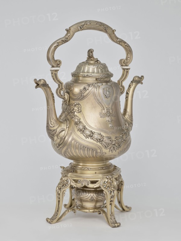 Kettle with warmer; maker: André Aucoc goldsmith; tableware, kettle; 1887–1911; France, Paris; vessel; height 39 cm, width 27 cm, depth 17 cm, A highly ornate metallic vessel with a tall curved handle and a hinged lid sits on an elaborately carved stand; the metal appears gold-toned and slightly aged, with intricate floral and scrollwork decorations covering the body, spout, handle, lid, and base. The vessel has a bulbous central body with raised decorative panels and garland motifs, a long curved spout with ornamental detailing at its tip, and a sculpted finial atop the lid. The handle is large and decorative with scroll shapes and carved accents. The stand is openwork with flowing, curved legs and matching ornamental motifs, and a small round container or burner is nestled beneath the vessel within the stand. The background is a plain light gray.