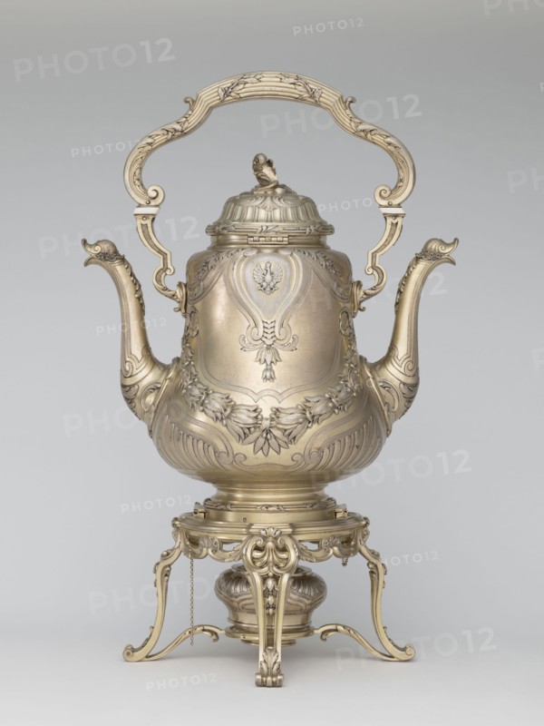 Kettle with warmer, by André Aucoc, goldsmith; tableware kettle, 1887–1911; France, Paris; vessel; height 39 cm, width 27 cm, depth 17 cm, A single ornate metal pitcher-like vessel with a large curved handle arching over its top, two elongated spouts on opposite sides, and a domed lid topped with a sculpted finial; the body and lid are decorated with embossed floral and foliate motifs, scrollwork, and carved cartouche shapes, and the vessel stands on a decorative tripod base with scrolling legs, a small suspended chain, and a round burner or reservoir beneath; visible colors include metallic gold tones, pale brass hues, and subtle shadows of gray against a neutral light gray background.