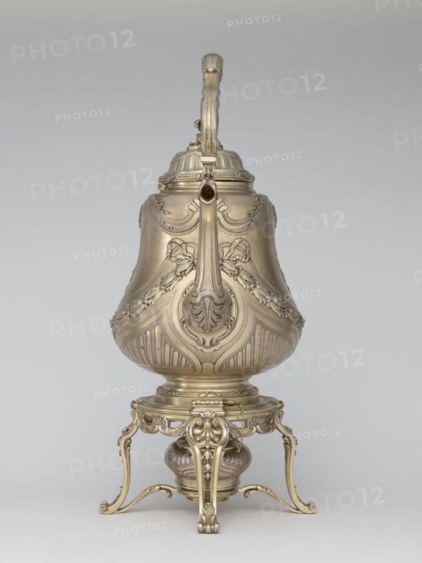 Kettle with warmer, maker: André Aucoc goldsmith, tableware item — kettle, 1887–1911, France, Paris; vessel; height 39 cm, width 27 cm, depth 17 cm, An ornate metallic vessel with a warm gold tone sits on an intricately carved stand of matching gold color; the vessel has a curved, bulbous body with embossed floral and ribbon motifs and fluted decorative panels, a hinged lid topped by a sculpted handle rising vertically, and a long spout extending from near the top; the stand features scrolling legs, acanthus-inspired carvings, and a rounded base element beneath the vessel; the background is a smooth, neutral gray.