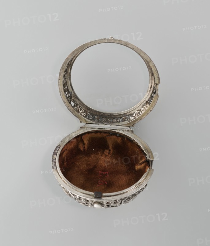 Pocket watch, Shart watchmaker. Pocket watch in a double silver case; inner case openwork with Regency motifs; outer case decorated with a repoussé scene depicting Diana hunting surrounded by an openwork border with male and female busts. White enamel dial with Roman numerals for hours and Arabic minutes, openwork hands. Signed on the movement: Shart / London. Date: 1730s, London, England, Great Britain. Timepiece; height 6.8 cm, width 4.8 cm., An open round metal compact with ornate carved edges reveals an inner lid with a circular mirror and an empty lower compartment lined with brown velvet; the metal is silver-toned with darker tarnish in places, the velvet lining is rich brown with a few slightly darker spots, and the background is a soft light gray.