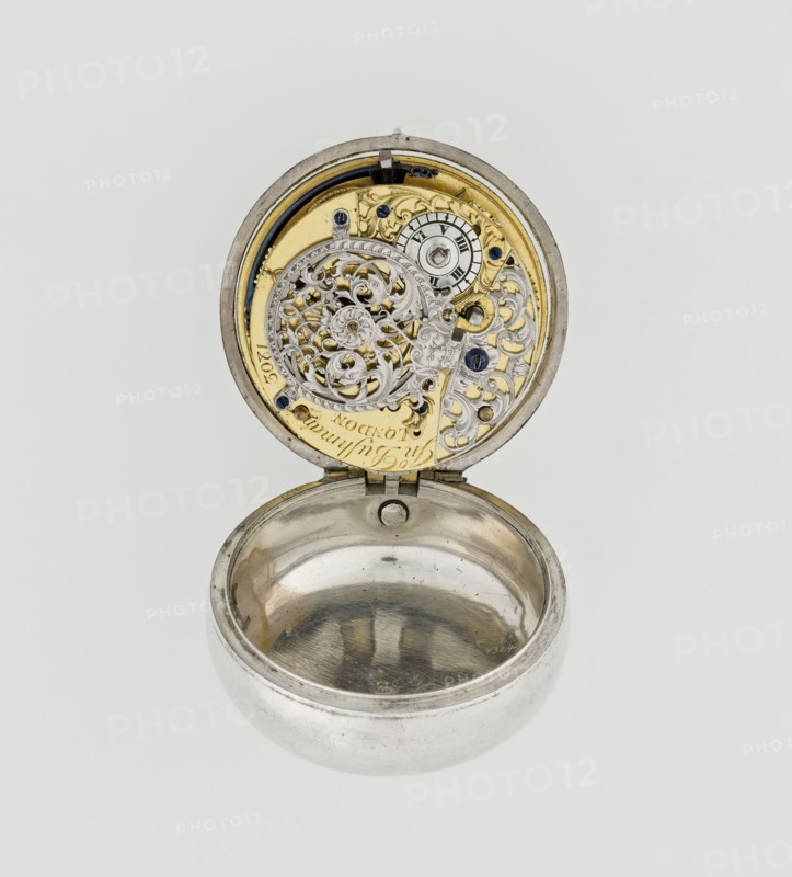 Pocket watch by John Bushman 1692–1720, c. 1700. Silver pocket watch in three cases: outer case covered with leather, rim decorated with studded silver rosettes; middle and inner cases plain. Underside of inner case stamped with casemaker’s initial H and number 3081. Transparent lid. Silver champlevé dial with Roman hour markers and Arabic minute markers in oval reserves on a matte background; central circular reserve with the watchmaker’s signature on two semicircular plates reading BUSHMAN and LONDON; date window at the bottom. Steel hands, openwork hour hand. Movement with plate skeleton, spring drive, worm gear, and Gall chain; spindle escapement. Gilded lower plate with an openwork English-style bun in the form of a wreath filled with acanthus tendrils and an openwork grid, connected by a mascaron mask with an openwork bun foot. Hair adjustment is a rotating silver-plated dial with scale I–VI. Engraved on the plate in italics: Jn Bushman 3081 / LONDON. Made in London, England. Tim...