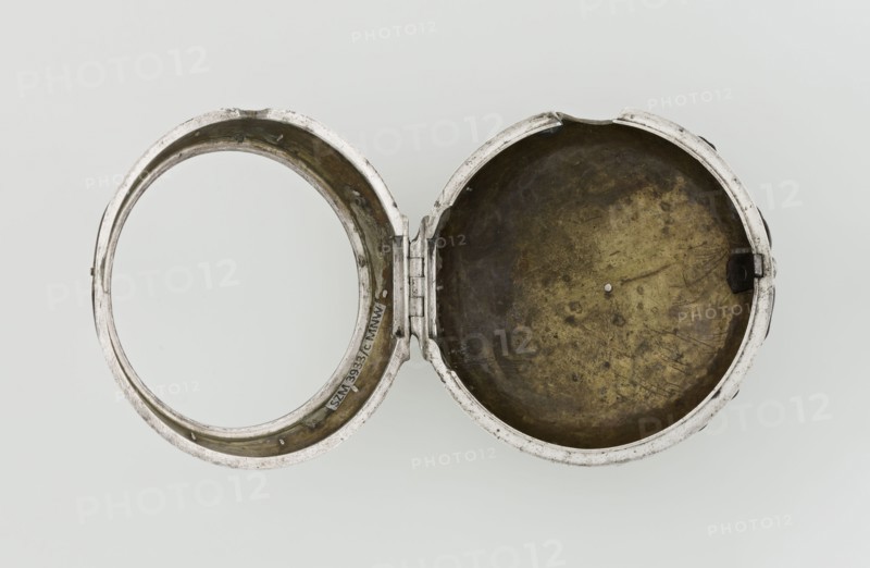 Pocket watch by John Bushman 1692–1720. Silver pocket watch in three cases: outer case covered with leather, rim decorated with studded silver rosettes, middle and inner cases plain; underside of the inner case stamped with the casemaker's initial H and the number 3081; transparent lid. Silver champlevé dial with Roman numeral hour markers and Arabic minute markers in oval reserves on a matte background; central circular reserve with the watchmaker's signature on two semicircular plates reading BUSHMAN and LONDON; date window at the bottom. Steel hands with openwork hour hand. Movement with plate skeleton, spring drive, worm gear, and Gall chain; spindle escapement. Gilded lower plate with an openwork English-style bun in the form of a wreath filled with acanthus tendrils and an openwork grid, connected by a mascaron mask with an openwork bun foot. Hair adjustment via a rotating silver-plated dial with a scale I–VI. Engraved on the plate in italics: Jn Bushman 3081 / LONDON. Circa 1700...