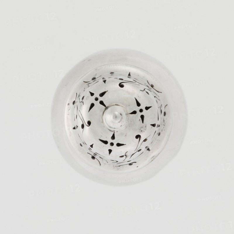 Powdered sugar bowl, powdered sugar bowl with domed openwork cover topped with a finial. Maker: Burbidge, Richard 1847–1917, goldsmith. Seller: Harrod's Stores Ltd London, fl. from 1849. Pear-shaped powdered sugar bowl on a circular profiled foot, with a domed openwork cover. Edge of the foot and the top rim decorated with astra gal ornament. Function: sugar vessel. Date: 1913–1914. Place: London, England, Great Britain. Materials/technique: openwork. Dish. Height 15 cm; diameter 5.9 cm; weight 152 g., A circular metallic object viewed from above with a domed center and a pattern of teardrop and curved cutouts arranged in a radial design, the surface showing subtle scratches and small dark spots; the predominant colors visible are silver, gray, and black, set against a pale off-white background.