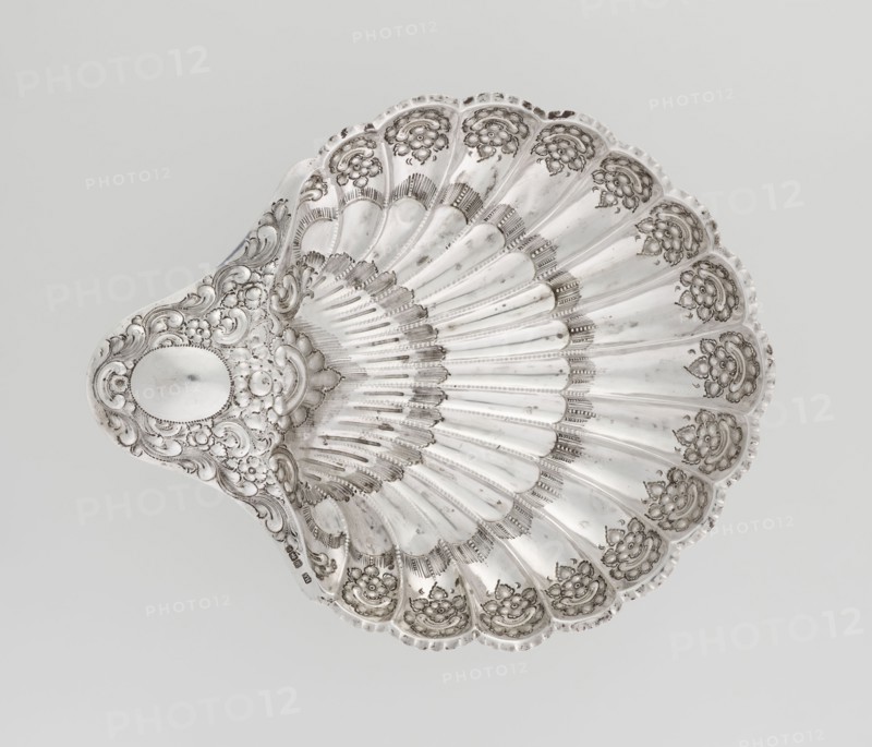 Shell-shaped silver platter by Atkin Brothers Silversmith Ltd Sheffield; goldsmith and silversmith firm; 1826–1958, scrap–purchase provenance; floral ornaments, scallop-shell shape, acanthus leaves ornament, clasp, relief decorations. Silver platter in the shape of a scallop shell, terminating on one side with an oval narrow handle, radial clasp, decorated on the rim with relief floral ornament, volutes and acanthus scrolls, supported on three shell feet. Dated 1893/1894, Sheffield, England. Vessel; height 5 cm, width 10 cm handle?, overall 28.5 × 23 cm; weight 357 g., A decorative shell-shaped dish in metallic silver tones fills the frame, showing a scalloped rim and radiating ridges that create a fan-like pattern; the surface features ornate raised floral and scrollwork embellishments in darker and lighter shades of silver, with areas of subtle shine and shadow that emphasize the embossed textures, and an oval smooth cartouche near the narrower end surrounded by intricate curling mot...