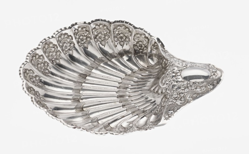 Shell-shaped silver platter by Atkin Brothers Silversmith Ltd Sheffield; goldsmith and silversmith firm; 1826–1958, 1893–1894. Scallop-shaped vessel terminating on one side with an oval, narrow handle; radially clasped; decorated on the rim with relief floral ornament, volutes and acanthus scrolls; supported on three shell feet. Materials: silver. Subject/ornament: floral ornaments, acanthus leaves, scallop shell motif. Dimensions: height 5 cm, depth 23 cm, width 28.5 cm, handle width 10 cm; weight 357 g., A decorative shell-shaped metal dish with a shiny silver surface, ornate raised floral and scrollwork patterns along the ridged interior and outer rim, a scalloped edge, and a smooth oval area near one end; colors visible include silver and shades of gray against a white background.