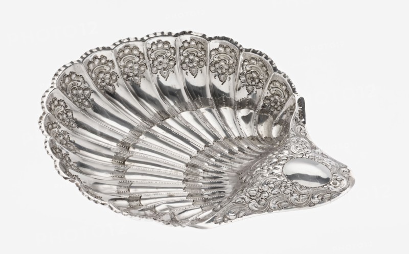 Shell-shaped silver platter by Atkin Brothers Silversmith Ltd Sheffield; goldsmith and silversmith firm; 1826–1958, 1893–1894. Silver platter in the shape of a scallop shell, terminating on one side with an oval narrow handle, radial clasp; rim decorated with relief floral ornament, volutes and acanthus scrolls; supported on three shell feet. Floral ornaments; acanthus leaves; scallop shape. Vessel. Height 5 cm; width handle? 10 cm; length 28.5 cm; depth 23 cm; weight 357 g. England, Sheffield., A decorative silver shell-shaped dish with a shiny metallic surface and ornate embossed floral and scrollwork patterns along the scalloped rim and the inner curved sections, showing a series of radiating ridges and grooves that follow the shell form; the metal reflects light creating areas of bright highlights and darker gray reflections, and the overall colors visible are silver and shades of gray against a white background.