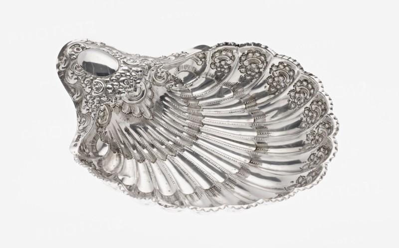 Shell-shaped silver platter by Atkin Brothers Silversmith Ltd Sheffield; goldsmith and silversmith firm; 1826–1958, 1893–1894. Scallop-shaped vessel terminating on one side with an oval narrow handle and radial clasp; decorated on the rim with relief floral ornament, volutes and acanthus scrolls; supported on three shell feet. Floral ornaments; acanthus leaves; relief decorations. Height 5 cm, depth 23 cm, width 28.5 cm, alternate width 10 cm, weight 357 g., A decorative shell-shaped metal dish with ornate raised floral and scrollwork detailing and a highly reflective silver surface, showing a few smooth, curved ridges radiating from the center and several scalloped edges, photographed against a plain white background with visible highlights and reflections.