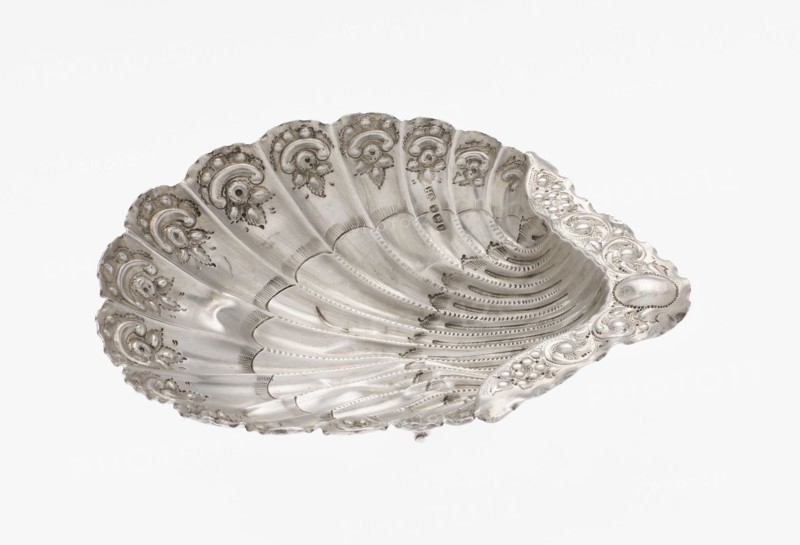 Platter in the form of a shell, Atkin Brothers Silversmith Ltd Sheffield; goldsmith and silversmith firm; 1826–1958 — Silver platter in the form of a full-plastic scallop shell, knotted and ribbed, decorated on the rim with a motif of relief flowers, leaves and channel sections; supported on three shell feet. Floral ornaments; shell shape; knotting; scallop shell. Date: 1890/1891. Place: Sheffield, England, Great Britain. Function: vessel. Dimensions: height 2.5 cm; width 15.3 cm; depth 13.1 cm; weight 113 g. Provenance: scrap — purchase, purchase., A metallic shell-shaped dish with a scalloped rim, displaying ornate raised floral and scrollwork decoration along the edges and interior; the surface shows fine engraved lines, dotted patterns, and textured bands radiating from the center, with areas of smooth polished metal and slight discoloration; overall color is silver with subtle darker gray accents where shadows and detailing appear.