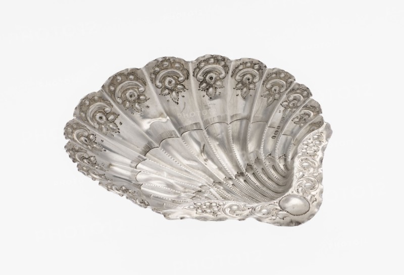 Platter in the form of a shell, Atkin Brothers Silversmith Ltd Sheffield; goldsmith and silversmith firm; 1826–1958 maker. Silver platter in the form of a full-plastic scallop shell, knotted and ribbed, decorated on the rim with a motif of relief flowers, leaves and channel sections; supported on three shell feet. Floral ornaments; shell shape; knotting; scallop shell. Vessel, 1890/1891, Sheffield, England, Great Britain. Height 2.5 cm; width 15.3 cm; depth 13.1 cm; weight 113 g., A decorative shell-shaped metal dish with a scalloped rim and a reflective silver surface, showing embossed floral and scroll motifs along the edges and in each fan-like segment; the interior has radiating ridges and textured dot patterns, with areas of subtle tarnish and darker gray shadows that emphasize the contours against a white background.