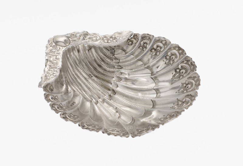 Platter in the form of a shell, Atkin Brothers Silversmith Ltd Sheffield; goldsmith and silversmith firm; 1826–1958 maker, scrap - purchase, floral ornaments, floral ornament, shell shape, knotting, scallop shell. Silver platter in the form of a full-plastic scallop shell, knotted and ribbed, decorated on the rim with a motif of relief flowers, leaves and channel sections; supported on three shell feet, 1890/1891, Sheffield, England, Great Britain. Vessel; height 2.5 cm, width 15.3 cm, depth 13.1 cm, weight 113 g., A scallop-shaped metallic dish with an ornate rim and a textured, ribbed interior, displaying decorative scrollwork and floral-like motifs along the edges; the surface is reflective with polished and matte areas, showing highlights and subtle shadowing; visible colors include silver tones, grey, and hints of darker tarnished grey.