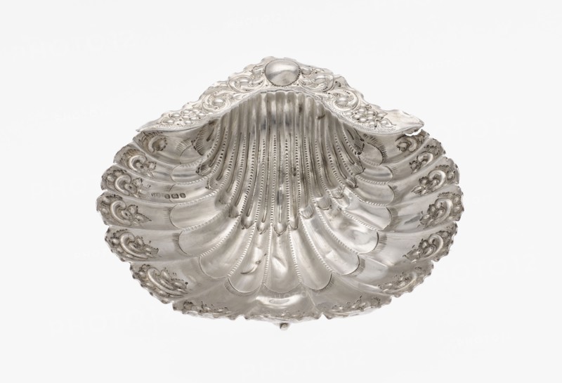 Platter in the form of a full-plastic scallop shell, knotted and ribbed, decorated on the rim with a motif of relief flowers and leaves and channel sections; supported on three shell feet. Maker: Atkin Brothers Silversmith Ltd Sheffield; goldsmith and silversmith firm; active 1826–1958. Date: 1890/1891. Place: Sheffield, England. Materials: silver. Function: vessel/platter. Dimensions: height 2.5 cm; width 15.3 cm; depth 13.1 cm; weight 113 g. Features: floral ornaments; shell shape; scallop shell; knotting; ribbing. Provenance: scrap — purchase., A single ornate shell-shaped object fills the frame, showing curved ridges and scalloped edges with embossed decorative motifs. The surface appears metallic with a polished sheen, reflecting light in gradients from bright highlights to softer gray tones. The object features repeated fan-like grooves radiating from the top center and small raised ornamental swirls along the rim. The overall colors visible are silver and varying shades of gray ...