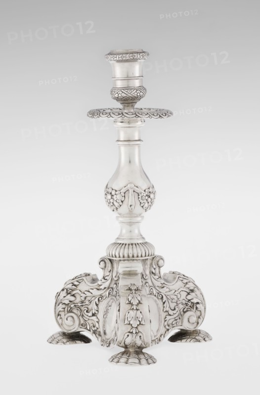 Single-candle candlestick, maker: Martin Goldstein fl. 1878–1886; maker firm: C.J. Vander, Ltd. London; goldsmith's firm, est. 1856; provenance: purchase scrap - purchase. Neo-Baroque style. Ornament: medallion, volutes, floral ornaments, godron ornaments, floral tendril. Description: single-candle candlestick in silver, supported on three shells and a three-sided raised base with side edges composed of volutes; richly decorated with floral tendril and plain medallions; baluster shaft at the bottom enclosed in a godron half-cylinder, decorated with floral festoons above, topped with a plate-shaped profit with a cylindrical sleeve. Date: 1884/1885. Place: London, England, Great Britain. Function: lighting. Dimensions: height 24 cm; width 13.6 cm; depth 13.6 cm; weight 852 g., A single ornate metallic candlestick stands upright against a light gray background, finished in a bright silver tone with darker areas where detailing creates contrast; the piece features a rounded top socket with...