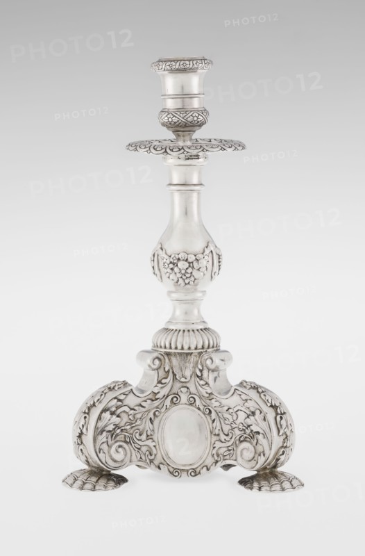 Single-candle candlestick by Martin Goldstein fl. 1878–1886 and the firm Vander, C.J., Ltd. London; goldsmith's firm, est. 1856. Neo-Baroque style; silver. Supported on three shells and a three-sided raised base with side edges composed of volutes, richly decorated with floral tendrils and plain medallions. Baluster shaft with bottom enclosed in a godron half-cylinder, decorated with floral festoons above, topped with a plate-shaped profiled element and a cylindrical sleeve. Ornamentation includes medallion, volutes, floral and godron ornaments. Dated 1884/1885, London, England. Function: lighting. Height 24 cm; width 13.6 cm; depth 13.6 cm; weight 852 g., A single ornate candlestick stands against a plain light background, made of a shiny metallic material with silver and gray tones; the candlestick has a flared holder at the top with decorative banding and a scalloped drip plate beneath it, a smooth tapered central column that transitions into a rounded midsection embellished with ra...