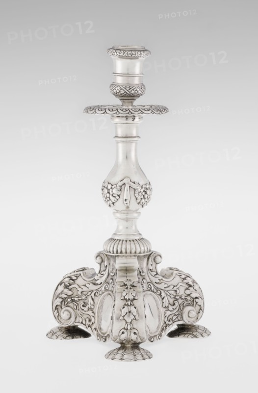 Single-candle candlestick by Martin Goldstein fl. 1878–1886 and Vander, C.J., Ltd. London; goldsmith's firm, est. 1856. Neo-Baroque style; silver. Supported on three shells and a three-sided raised base with side edges composed of volutes, richly decorated with floral tendrils and plain medallions. Baluster shaft at the bottom enclosed in a godron half-cylinder, decorated with floral festoons above, topped with a plate-shaped drip pan and a cylindrical sleeve. Medallion, volutes, floral and godron ornaments. Date: 1884/1885. Place: London, England, Great Britain. Function: lighting. Dimensions: height 24 cm; width 13.6 cm; depth 13.6 cm. Weight: 852 g., A single ornate metal candlestick stands against a plain light background, showing a polished silver surface with darker toned recesses that highlight intricate carving and relief work; the base flares into a decorative tri-lobed platform with a mix of scrolls, acanthus-like leaves, shell-shaped feet, and raised floral motifs, while the...