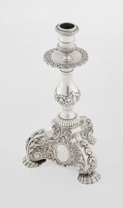Single-candle candlestick, neo-Baroque style, silver, supported on three shells and a three-sided raised base with side edges composed of volutes, richly decorated with floral tendril and plain medallions; baluster shaft at the bottom enclosed in a godron half-cylinder, decorated with floral festoons above, topped with a plate-shaped profit, with a cylindrical sleeve. Medallion, volutes, floral ornaments, godron ornaments, floral tendril. Date: 1884–1885, London, England Great Britain. Function: lighting. Dimensions: height 24 cm; width 13.6 cm; depth 13.6 cm; weight 852 g., A silver-colored ornate candlestick with a decorative circular cup at the top, a flared scalloped drip tray beneath it, and a turned column that narrows then widens into a bulbous midsection adorned with raised floral garlands; the base is an elaborately carved three-footed platform with scrolling acanthus motifs, shell-like feet, and oval cartouche panels, all rendered in metallic silver and subtle darker patina a...