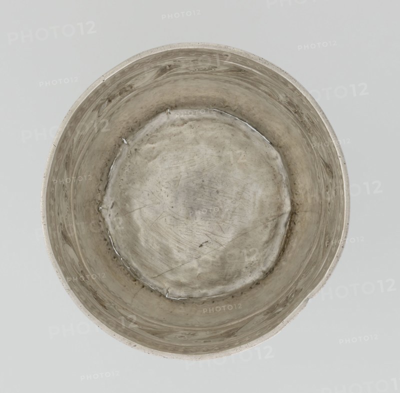 Bowl by William I Sumner fl. 1773–ca. 1807, goldsmith. Circular, bulging vessel with a flat bottom and flared upper edge; decorated on the belly with a wide band of relief ornament of acanthus leaves and flowers. Materials: metal vessel. Styles: Georgian, George III, Empire; motifs: acanthus. Date: 1804–1805. Place: London, England. Dimensions: height 8 cm; diameter 11.5 cm; weight 145 g. Provenance unknown., A top-down view of a round metal container with a dull silver-gray surface showing irregular scratches, scuffs, and areas of darker tarnish; the interior rim and walls display subtle swirling patterns and a few small raised seams, and the bottom appears slightly concave with uneven texture and faint shadowing; the background is a flat pale gray.