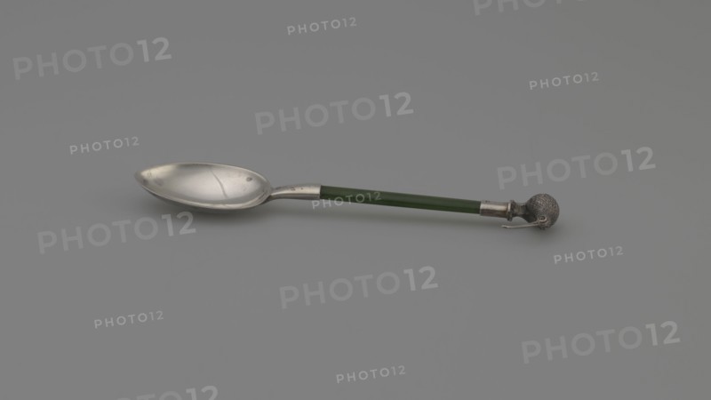 Souvenir spoon by FH goldsmith; Birmingham; fl. ca. 1903–1904. Souvenir spoon with an oval scoop, a flat jade handle set in a silver sleeve, and topped with a silver full-plastic kiwi bird figurine. Likely made in Birmingham, England, ca. 1903–1904. Cutlery; height 10.8 cm, width 2.3 cm., A single spoon lies horizontally on a plain background, showing a metallic bowl with a smooth, slightly reflective surface and some small marks, a straight handle that is green and cylindrical, and a decorative metallic end shaped like a textured sphere with a small protruding element; the visible colors include silver, green, and shades of gray in the background.