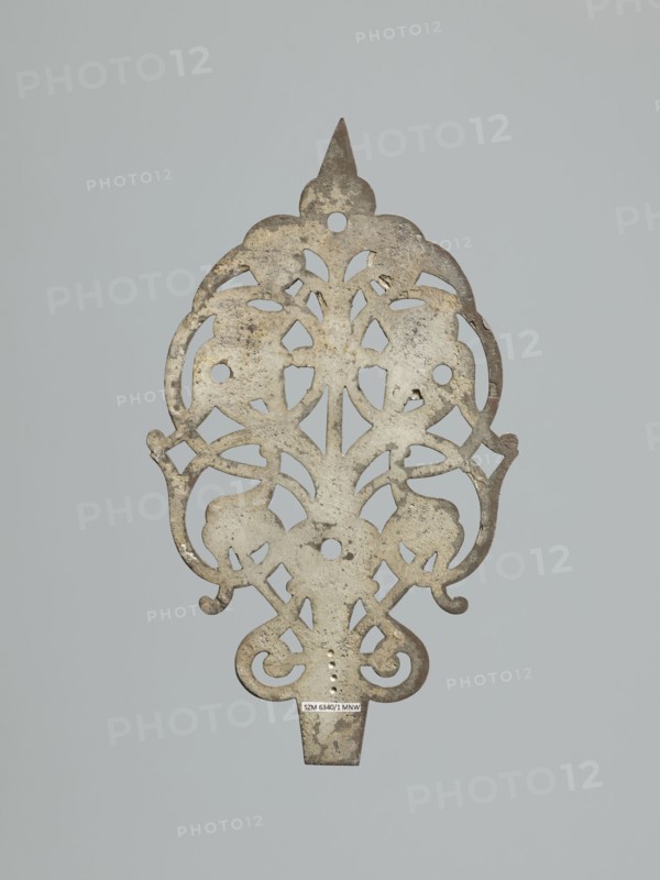 Hinge end for a chest; fittings, hinge end from a pair, in the form of an openwork rounded plaque with a tendril motif, two male masks among the twists. Hinge cut off; perhaps originally a disc hinge with a sleeve at the end of the plate or a strap hinge in the form of a clock hand. 17th century, Poland, Central Europe. Door fittings. Height 21.5 cm, width 12.5 cm, depth 0.2 cm. Maker: Roman Szewczykowski 1849–1901, An ornate, flat metal object with a symmetrical, openwork design featuring curving, scroll-like shapes and floral or leaf-like cutouts, showing a weathered surface with patches of darker and lighter areas; the metal appears mainly gray with areas of brownish tarnish and pale off-white spots, set against a uniform light gray background.