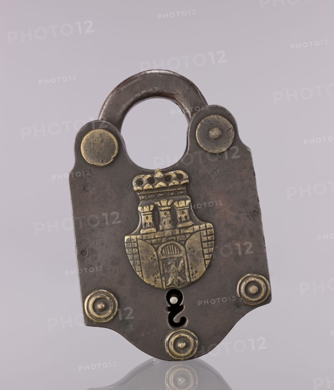 Padlock with the coat of arms of Kraków and brass rosettes, coats of arms, late 18th–early 19th century, Kraków, Poland; locksmithing; height 19 cm, width 12.5 cm, A metal padlock with a rounded shackle and a flat, shield-shaped body sits upright against a pale lavender-gray background. The lock shows a dark brown to blackened patina with areas of worn brass-colored metal accents, including a decorative brass emblem in the center featuring a sculpted architectural motif and a few circular brass rivets positioned near the top and bottom edges. The keyhole area is visible as a dark opening near the lower center, and the surface of the lock displays subtle scratches and mottled discoloration in shades of brown and dark gray. The reflection of the lock appears faintly at the bottom on a glossy surface.