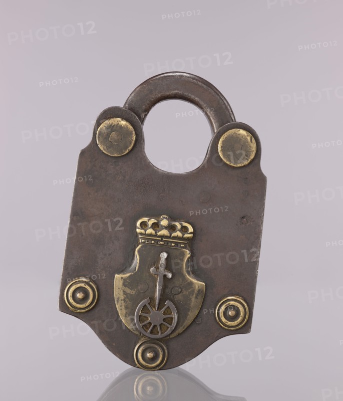 Padlock with the coat of arms of Kraków and brass rosettes; maker, coats of arms; late 18th–early 19th century; Poland, Kraków ; locksmithing; height 19 cm, width 12.5 cm., A metal padlock with a rounded shackle and a broad, flat body sits against a pale mauve background; the lock body is dark brown with a slightly mottled, aged surface and has several brass-colored decorative elements including round rivet-like discs near the top and bottom, a brass shield-shaped ornament in the center topped by a small crown-like piece, and a wheel-and-cross motif on the shield; the brass accents show a yellowish-gold tone contrasted with the darker brown metal, and the lock casts a faint reflection on a glossy surface below.