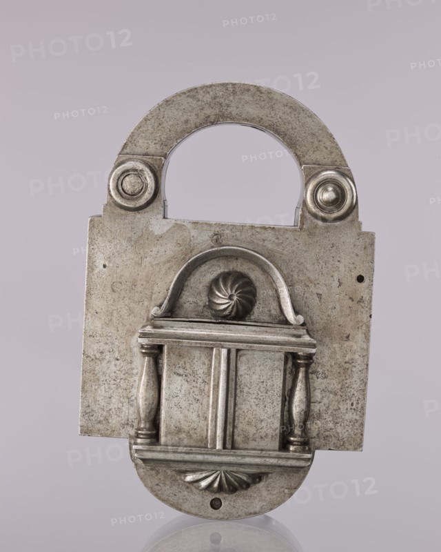 Rectangular padlock with a bow, 19th century, Polesie; locksmithing; height 18.3 cm, width 11.7 cm, depth 5.4 cm., A metallic padlock with a weathered silver-gray surface stands against a pale lavender-gray background, showing an arched shackle with round rivet-like details at its base, a rectangular main body with a raised decorative front panel featuring an arched top, a central spiral motif, two turned column-like elements flanking a recessed central section, and a rounded lower flap with a shell-like motif, the metal showing areas of darker tarnish and lighter wear.
