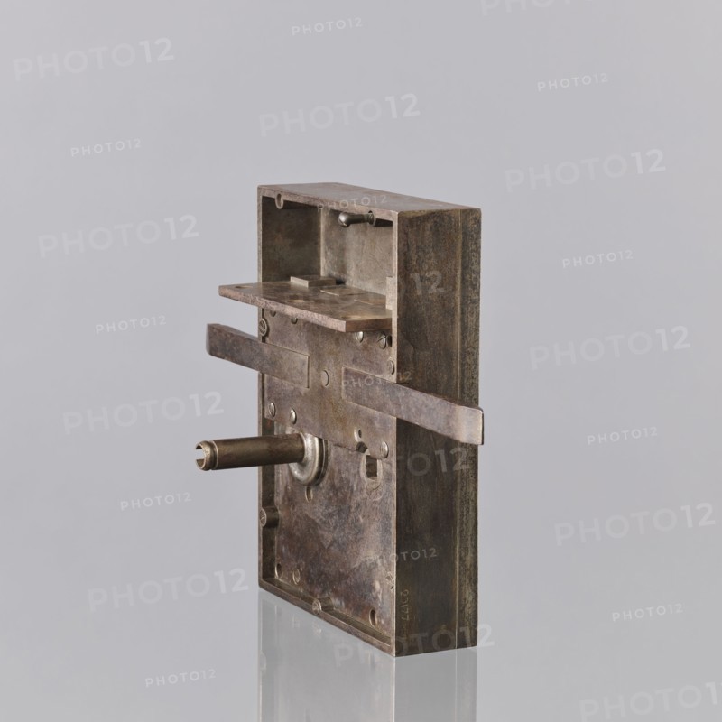 Vault lock by Karol Konstabel, locksmith, 2nd half of the 19th century, Warsaw. Masterpiece marked with the guild stamp stamp now invisible. Rectangular cup with an overlaid border with inner wavy edge, a three-length vertical strip with a sequin and two inverted lyre shapes. The lock has three bolts and a fourth latch bolt connected to the handle handle missing. On the underside the case with the mechanism is separated, with a narrow chamber that would cover the bolt hasps. The workmanship and the key register are among the more outstanding features. Same technical solution as in a handmade lock described as Taszynski's from 1861; typical design for a main-door treasury lock with flanking riffles, two springing and two lifting fels. Height 40 cm, width 27.5 cm, depth 12.5 cm. Karol Konstabel was probably the son of Wilhelm, a master locksmith., A metal rectangular mechanism with a weathered brown and dark gray patina stands upright against a pale gray background, showing a hollow top ...