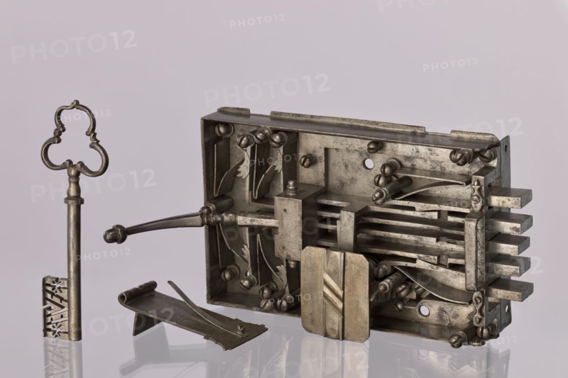 Gate lock. Double-bolt gate lock with key No. 52. Uncapped, with pull handle. Housed in a rectangular casing, with five bolts: three central bolts slide back and forth; the two outermost are scissor-type which, when opened, lock the other bolts via side projections engaging cut-out recesses. Unlocking achieved by lifting the lever at the end of the lock or by turning the key. Key register box closed with a rectangular plate with cut corners and a central strip with oblique cuts. Very good, clean workmanship; indicative of dedicated earlier craftsmen. 17th/18th century, possibly Austria; locksmith, Height 17.5 cm, width 35 cm., A metallic lock mechanism and key rest on a reflective surface against a pale background; the lock body is rectangular and open to reveal internal components including several flat bars, curved levers, screws, and a sliding block, all in a silvery metal with areas of darker tarnish and patina, while the detached key stands upright with an ornate bow and a long sh...