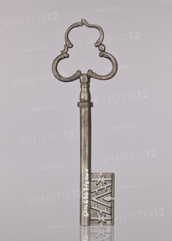 Gate key, locksmithing. Key with a tubular shaft, separated from the semi-ovoidal node by a flat cylinder; handle made of three C-sections. The blade widens downward, with a rib, diagonal notches and two stars. 17th/18th century, possibly Austria; height 19.8 cm, width 6.2 cm, depth 1.3 cm., A single ornate metal key is centered against a pale gray background, with a decorative clover-shaped bow featuring curved, stylized cutouts and a smooth cylindrical shaft leading to a rectangular bit that has intricate angular cut patterns; the key appears metallic with tones of silver and pewter, showing slight variations in darker and lighter gray, and it casts a faint reflection on the glossy surface below.